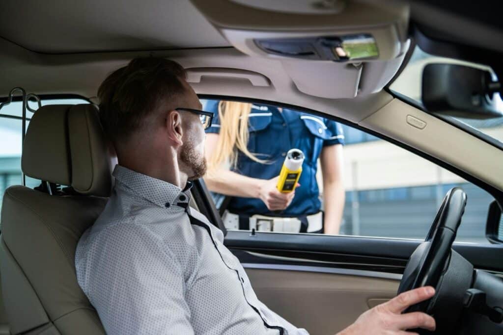 A driver seated in a car while a police officer holds a breathalyzer device outside the window