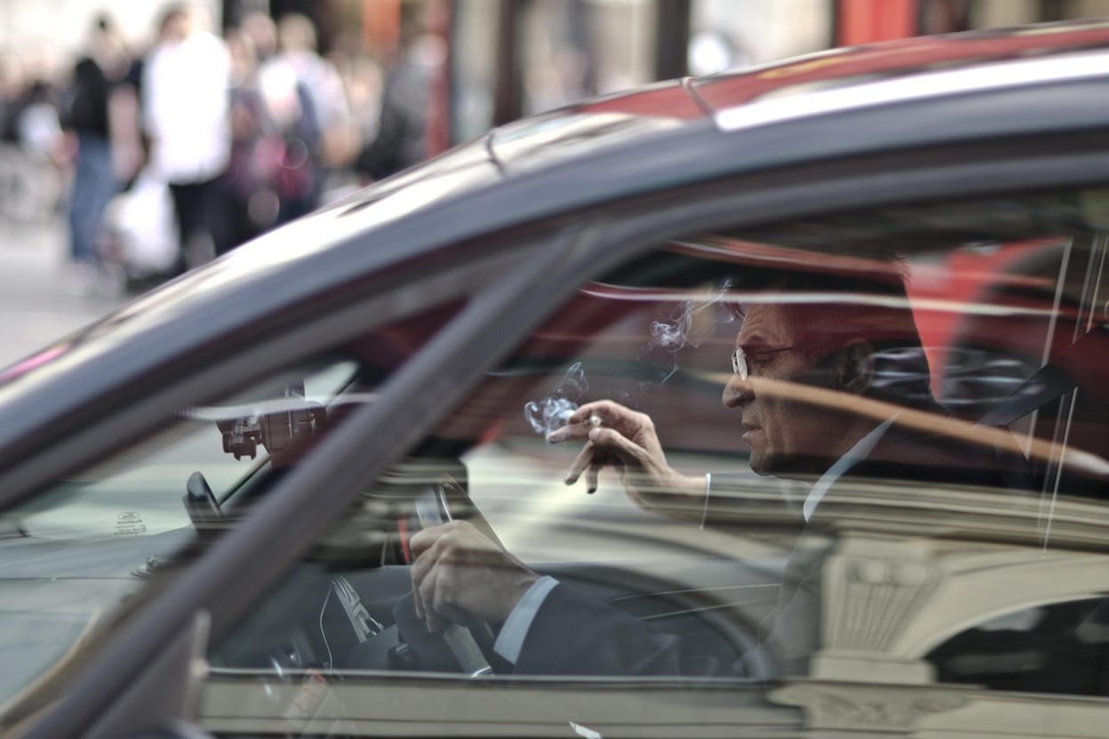 A man in a suit smoking a cigarette while sitting behind the wheel of a car