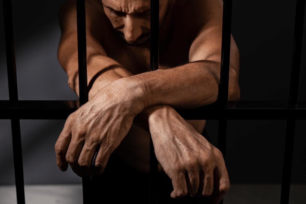 A man sitting behind prison bars with his arms resting through the bars