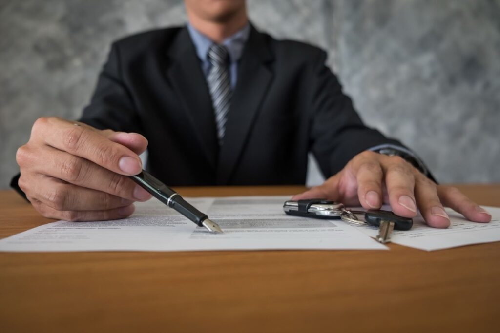 A suited man points a pen at a legal document with car keys on the table