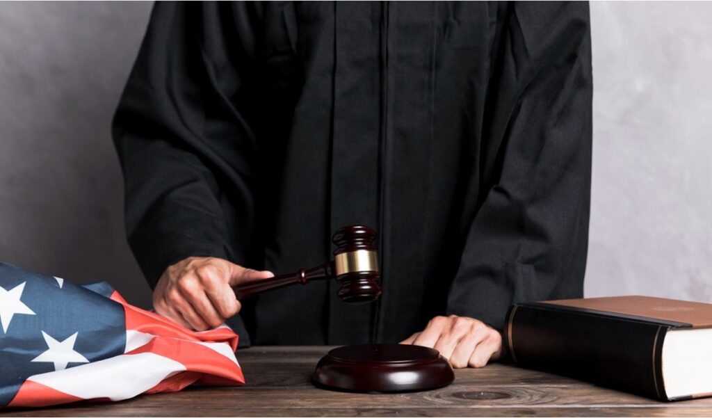 Judge in a black robe holding a gavel over a sound block with an American flag on the bench