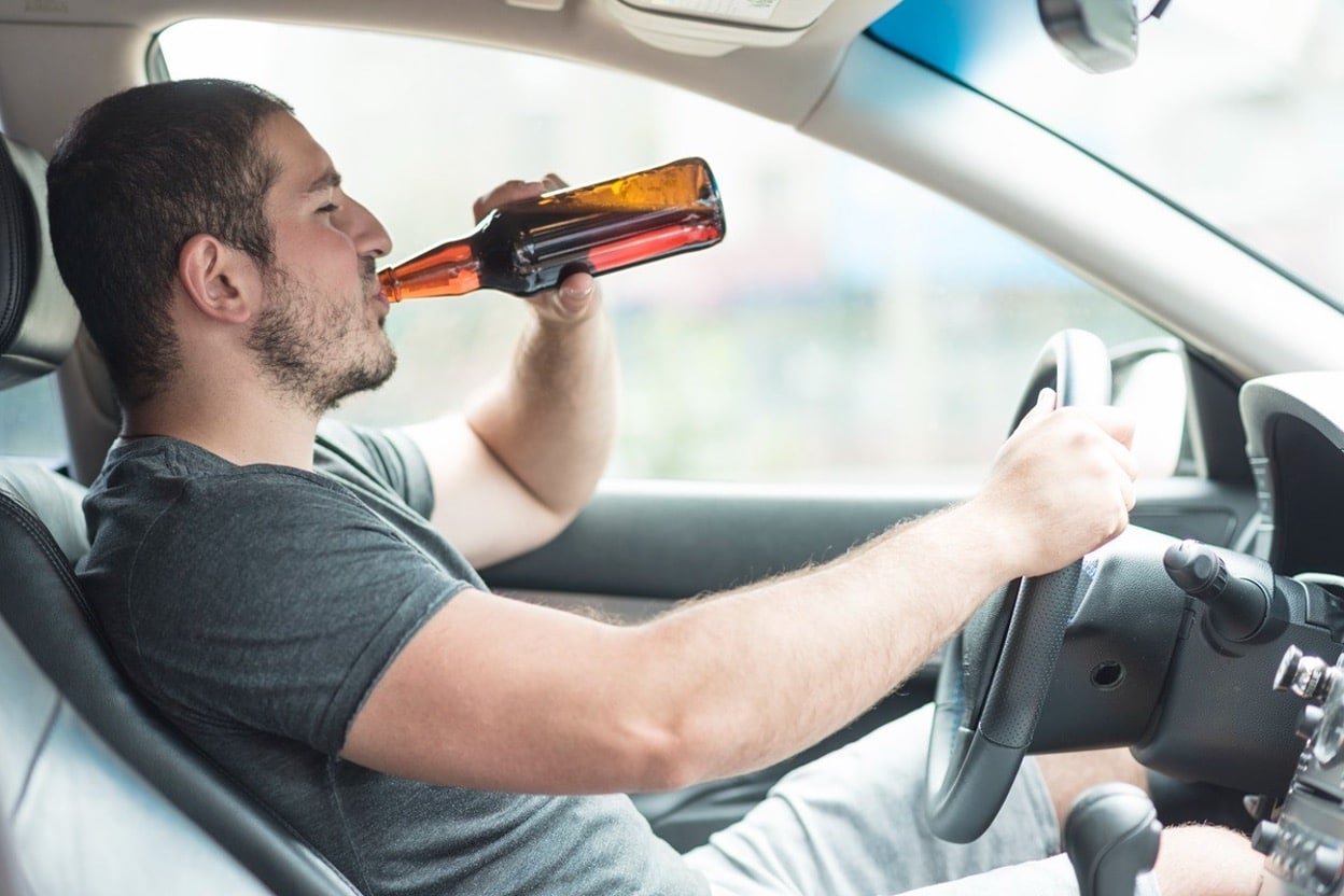 Man drinking from a beer bottle while sitting behind the steering wheel of a car