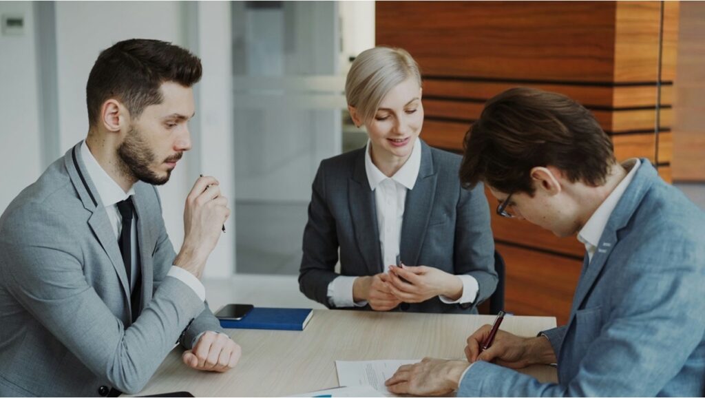 Three people seated at a table reviewing and signing documents in an office setting