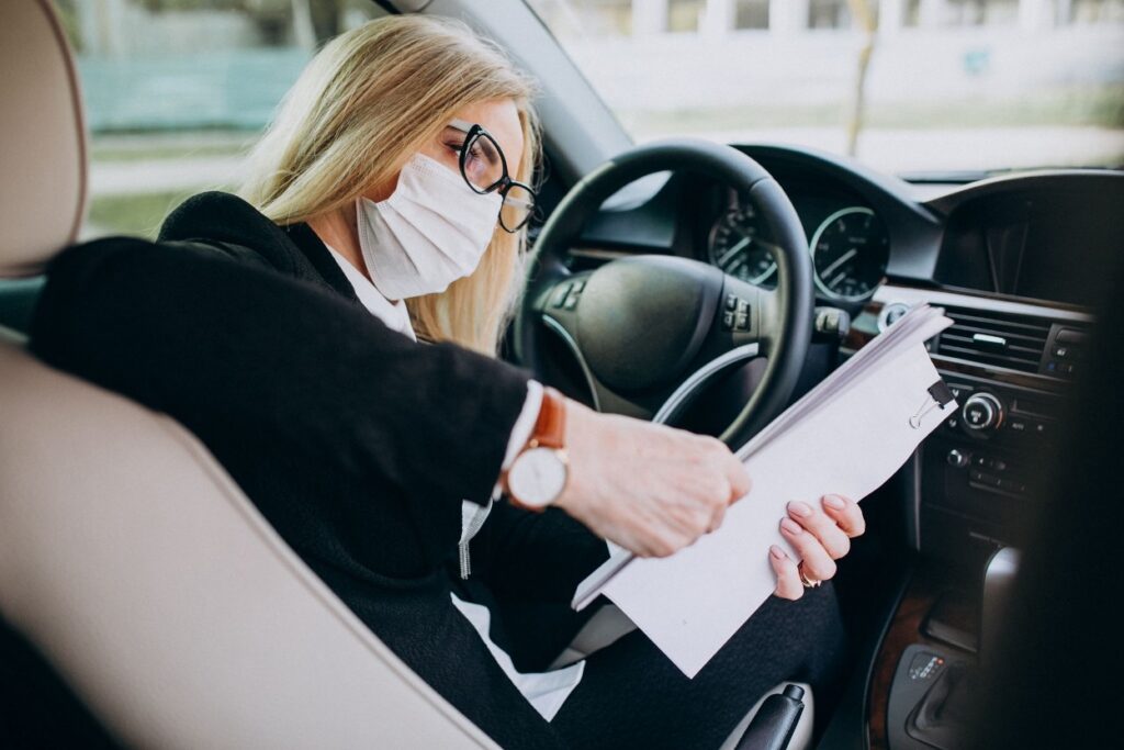 A blonde woman wearing a face mask and glasses sits in the driver's seat of a car, reading papers