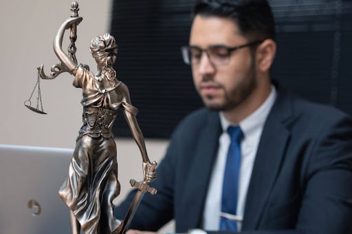 A bronze Lady Justice statue in the foreground with a lawyer working on a laptop in the background.