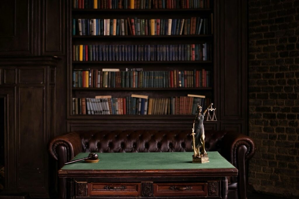 A dark wood-paneled law library featuring a green-topped desk with a judge's gavel and a bronze Lady Justice statue