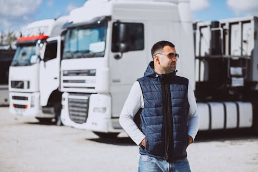 A male truck driver standing confidently in front of a fleet of white semi-trucks.