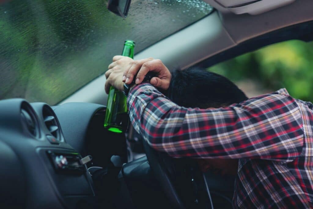 A man slumped over a car's steering wheel while holding a green beer bottle, depicting drunk driving