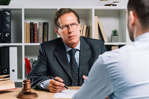 A senior lawyer in a suit listening attentively to a client across the desk in a professional office.