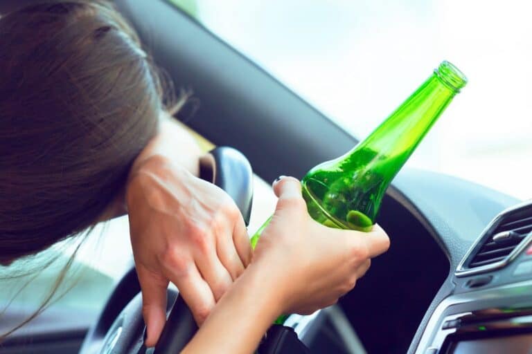 A woman slumped over the steering wheel, holding an open beer bottle while sitting in the driver's seat.
