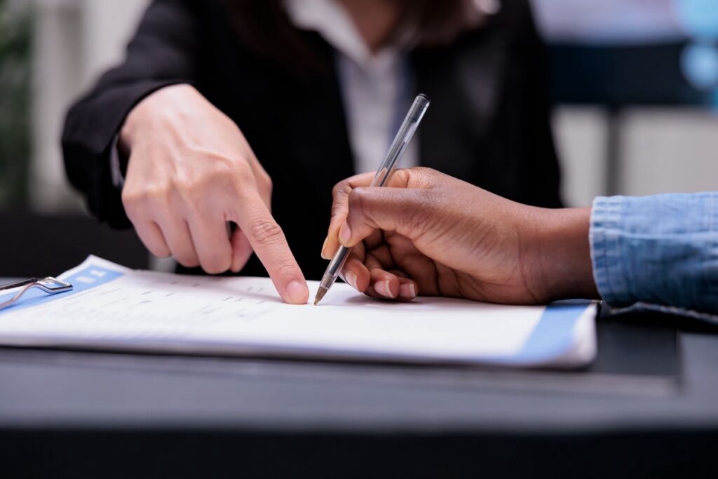 Two people's hands over a clipboard, with one pointing to a form and the other signing with a pen